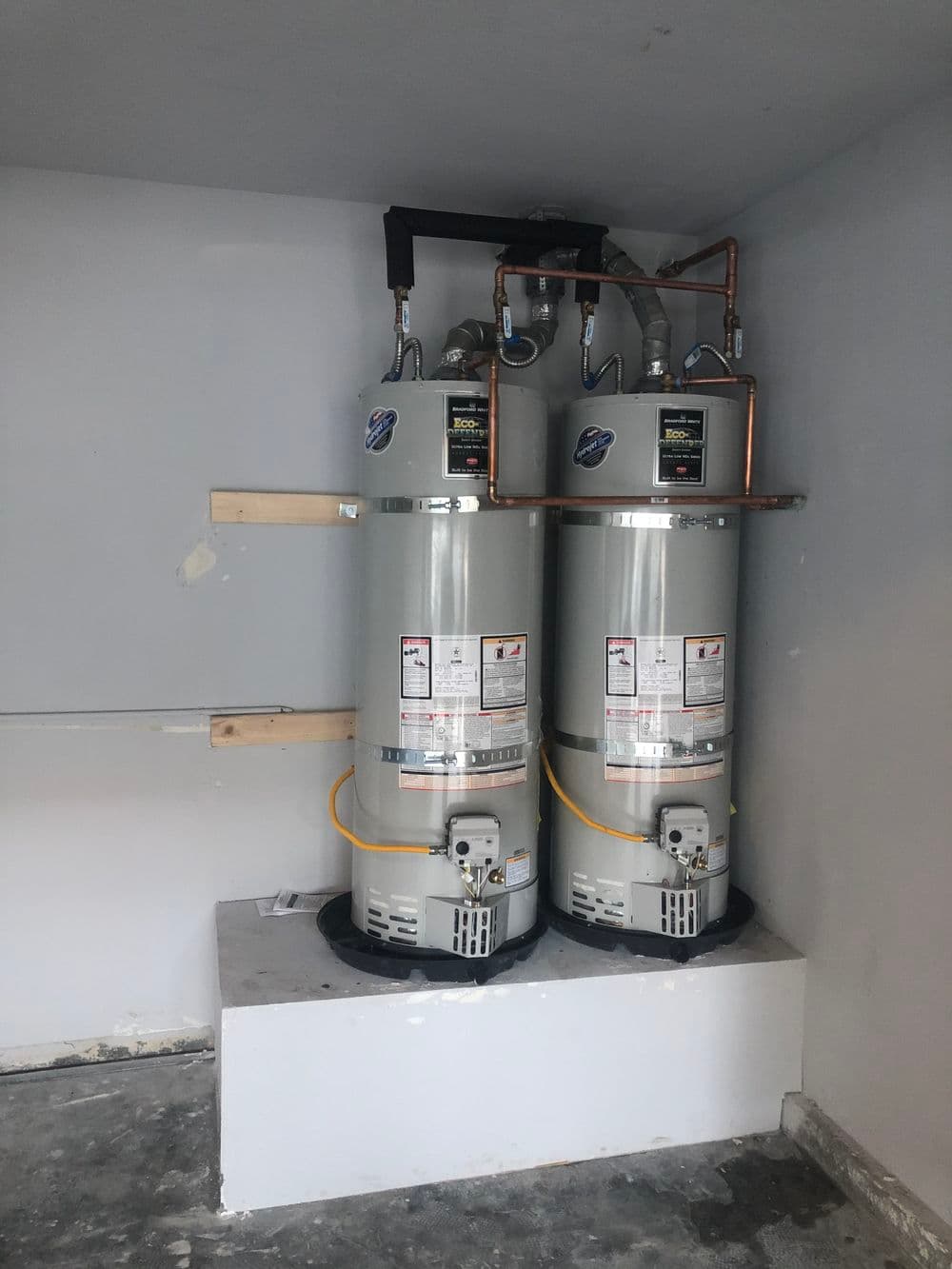 Two vertical water heaters installed side by side in a utility room with exposed pipes.