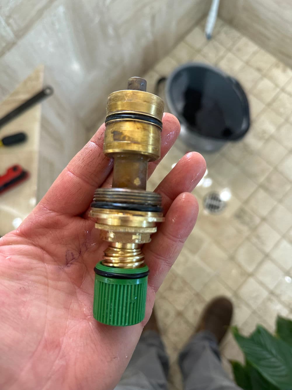 Hand holding a brass shower valve repair part with a green seal, set against a tiled background.