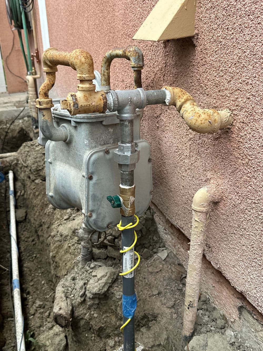 Natural gas meter with rusted pipes installed on residential wall near a buried line.