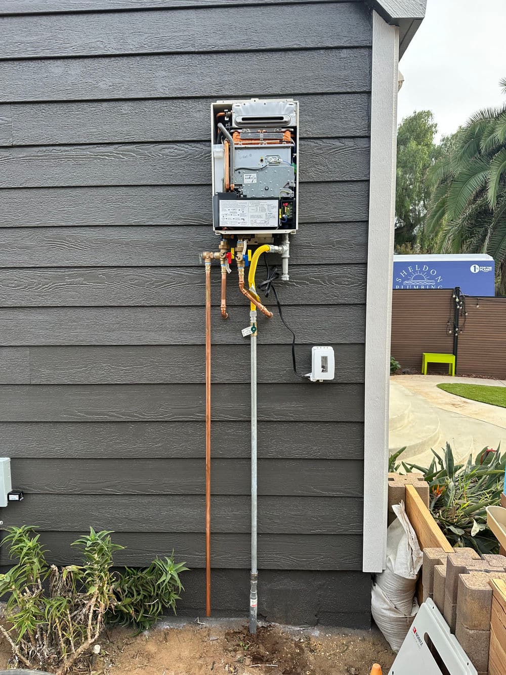 Tankless water heater installation on a dark gray wall with plumbing connections and nearby landscaping.